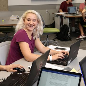 A student smiles while glancing at the computer screen of another student.